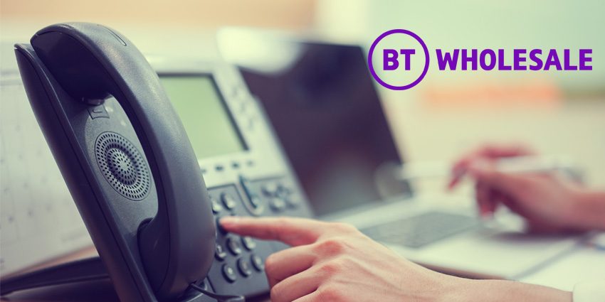 Man's hand at foreground hitting button on landline phone while other rests in blurred background against laptop and BT Wholesale logo above