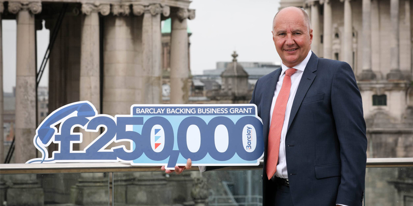 Barclay Communications Founder Britt Megahey standing aginst sandstone buildings with sign saying £250000