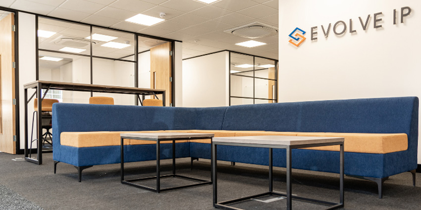 Wooden coffee table surrounded by blue L-shaped couch and tall table in background in beige office with Evovle IP logo on wall