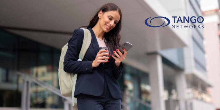 A business professional walking outside an office building, smiling while checking her smartphone and holding a coffee cup with Tango Networks logo in the top right corner